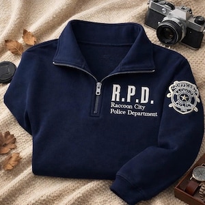 Op de afbeelding: Marineblauwe trui met een kwart rits, met de tekst "R.P.D. Raccoon City Police Department" en een politielogo op de mouw. Een vintage camera en horloge zijn zichtbaar.
