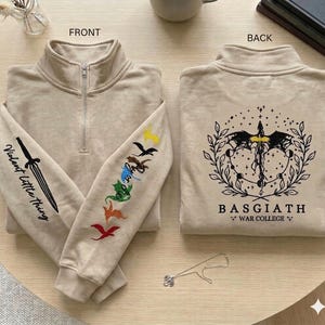 May include: Beige quarter-zip sweatshirt with embroidered details. The left sleeve features a sword and the text "Violent Little Thing." The right sleeve has colourful dragon designs. The back displays a graphic with the text "BASGIATH WAR COLLEGE."