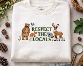May include: White sweatshirt with an embroidered design featuring a bear, fox, bird, raccoon, and deer. The phrase "Respect the Locals" is embroidered in green. The sweatshirt is folded and displayed on a white surface.