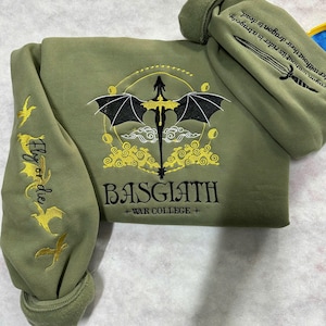 May include: A green sweatshirt with a black dragon and yellow clouds embroidered on the front. The text "Fly or die" is embroidered on the left sleeve. The text "BASGIATH +WAR COLLEGE+" is embroidered below the dragon.