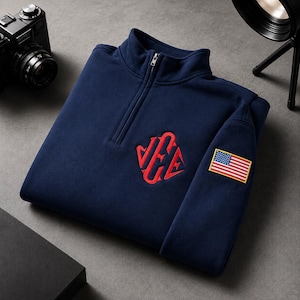 May include: A navy blue quarter-zip pullover featuring a red embroidered monogram on the chest and a small rectangular patch of the United States flag on the sleeve. The garment has a high collar with a silver-toned metal zipper closure.