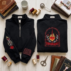 May include: Black quarter-zip sweatshirt with embroidered designs. The front has a silver zip and the left sleeve features a red and gold celestial design. The back displays a red and gold heart and sword design with the text "FIREHEART".