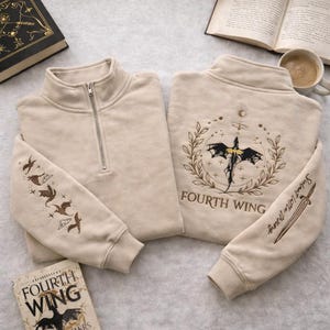 Può includere: Felpa beige con zip a un quarto con ricami di draghi e spade. Il retro della felpa presenta la scritta "FOURTH WING" e un'emblema di drago. La manica ha la scritta "Sgaeyl Little Wing". È visibile anche un libro intitolato "FOURTH WING".