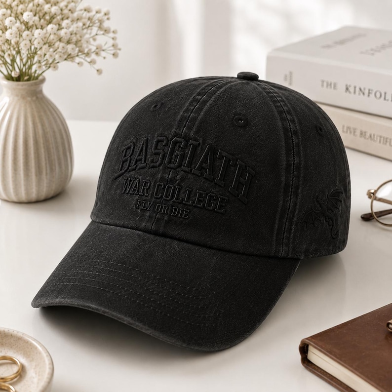 May include: Black baseball cap with the text "BASCIATH WAR COLLEGE FLY OR DIE" embroidered on the front. The cap has a curved brim and a small emblem on the side. The cap is sitting on a white surface.
