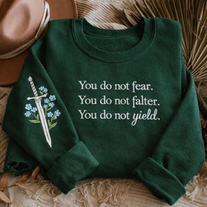 May include: A dark green sweatshirt with the text "You do not fear. You do not falter. You do not yield." in white. The left sleeve features an embroidered sword and blue flowers.