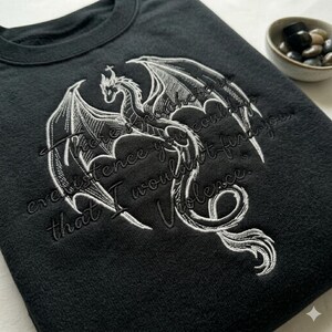 Puede incluir: Sudadera negra con un dise&ntilde;o de drag&oacute;n bordado en blanco y la cita: "Donde la existencia no te encontrar&iacute;a, yo encontrar&iacute;a la violencia". El drag&oacute;n tiene alas detalladas.