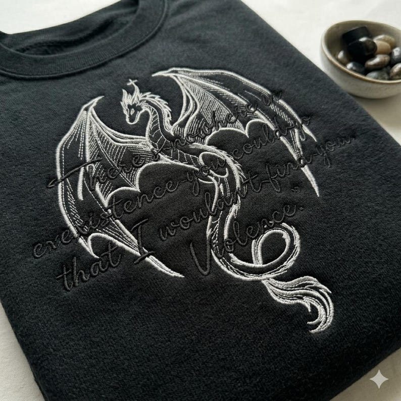 Puede incluir: Sudadera negra con un dise&ntilde;o de drag&oacute;n bordado en blanco y la cita: "Donde la existencia no te encontrar&iacute;a, yo encontrar&iacute;a la violencia". El drag&oacute;n tiene alas detalladas.