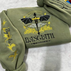 May include: A green sweatshirt with a black dragon and gold embroidery. The text "Fly or die" is embroidered on the sleeve. The text "BASGIATH +WAR COLLEGE+" is embroidered on the front.