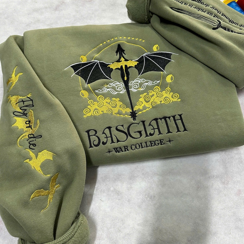 May include: A green sweatshirt with a black dragon and gold embroidery. The text "Fly or die" is embroidered on the sleeve. The text "BASGIATH +WAR COLLEGE+" is embroidered on the front.