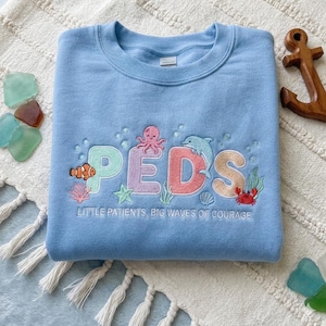 Op de afbeelding: Een lichtblauwe sweatshirt met het geborduurde woord "PEDS" in kleurrijke letters, met een octopus, dolfijn en andere zeedieren. De tekst "LITTLE PATIENTS, BIG WAVES OF COURAGE" staat eronder geborduurd.