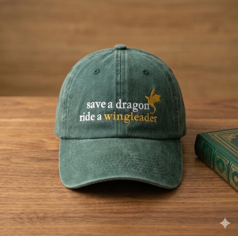 May include: A green baseball cap with the embroidered text "save a dragon ride a wingleader" in white and gold. The cap features a curved brim and a six-panel design. The cap is sitting on a wooden surface.