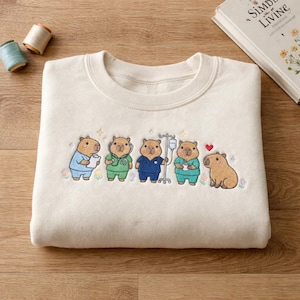May include: Cream-coloured sweatshirt with an embroidered design of capybaras dressed as medical professionals. The capybaras wear scrubs and hold medical equipment. A red heart is embroidered next to the last capybara. A book and spools of thread are in the background.