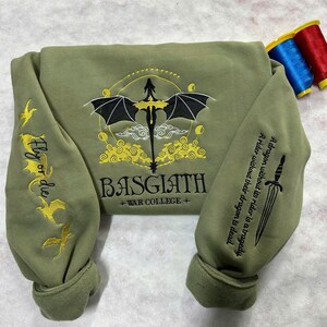 May include: A green sweatshirt with a black dragon embroidered on the front. The dragon is surrounded by yellow clouds and the text "BASGIATH +WAR COLLEGE+". The sleeves have yellow embroidered dragons and the text "Fly or die".