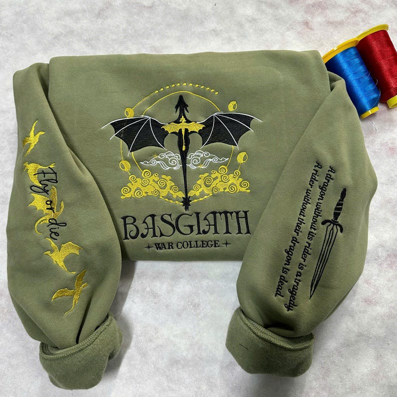 May include: A green sweatshirt with a black dragon embroidered on the front. The dragon is surrounded by yellow clouds and the text "BASGIATH +WAR COLLEGE+". The sleeves have yellow embroidered dragons and the text "Fly or die".