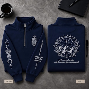 Könnte beinhalten: Ein marineblaues Sweatshirt mit Viertelreißverschluss und weißen Linienzeichnungen. Die Vorderseite zeigt eine Kapuzenfigur, Mondphasen und den Text "Only you can decide what breaks you". Die Rückseite zeigt eine Berglandschaft, Sterne und "VELARIS CITY OF STARLIGHT to the stars who listen and the dreams that are answered."