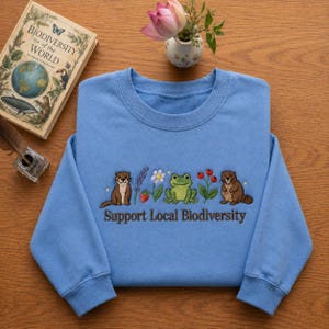 Peut inclure: Un sweat-shirt bleu clair avec un motif brodé représentant une loutre, de la lavande, une fraise, une grenouille, des fleurs, des baies et un castor. Le texte "Support Local Biodiversity" est brodé en dessous. Un livre et un vase de fleurs sont en arrière-plan.