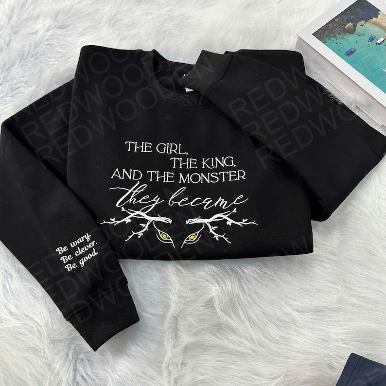 Peut inclure: Sweat-shirt noir avec l'inscription blanche : "THE GIRL, THE KING, AND THE MONSTER they became." En dessous, deux yeux jaunes stylis&eacute;s. La manche gauche porte l'inscription "Be wary. Be clever. Be good."
