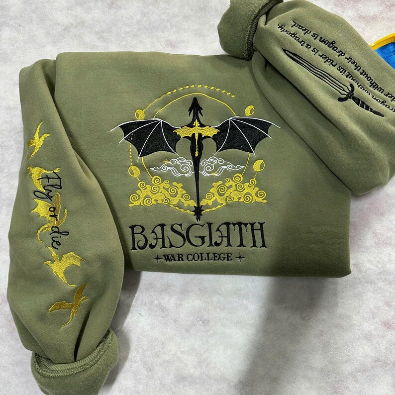 May include: A green hooded sweatshirt with a black dragon and yellow clouds embroidered on the front. The text "BASGIATH +WAR COLLEGE+" is embroidered below the dragon. The left sleeve has the text "Fly or die" embroidered in yellow.