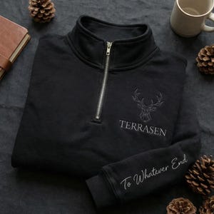 Peut inclure: Sweat-shirt noir à fermeture éclair quart de zip argentée. Il présente un motif de tête de cerf géométrique et le mot "TERRASEN" brodé en blanc. La manche porte l'inscription "To Whatever End" en écriture blanche.