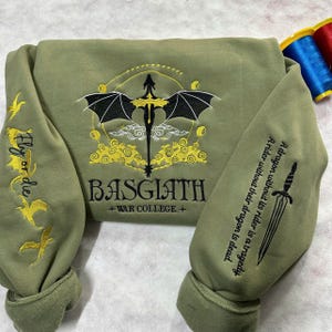 May include: A green sweatshirt with a black dragon embroidered on the front. The dragon is surrounded by yellow clouds and the text "BASGIATH +WAR COLLEGE+". The sleeves have embroidered text that reads "Fly or die" and "A dragon without its rider is a tragedy. A rider without their dragon is dead."