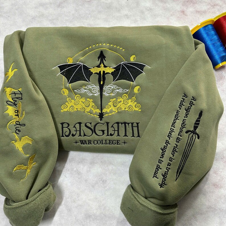 May include: A green sweatshirt with a black dragon embroidered on the front. The dragon is surrounded by yellow clouds and the text "BASGIATH +WAR COLLEGE+". The sleeves have embroidered text that reads "Fly or die" and "A dragon without its rider is a tragedy. A rider without their dragon is dead."