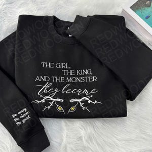 Peut inclure: Sweat-shirt noir avec l'inscription blanche "THE GIRL, THE KING, AND THE MONSTER they became". Le design comprend des yeux stylis&eacute;s et des branches. La manche porte l'inscription "Be wary. Be clever. Be good."
