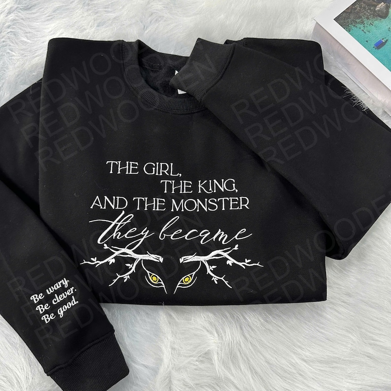Peut inclure: Sweat-shirt noir avec l'inscription blanche "THE GIRL, THE KING, AND THE MONSTER they became". Le design comprend des yeux stylis&eacute;s et des branches. La manche porte l'inscription "Be wary. Be clever. Be good."