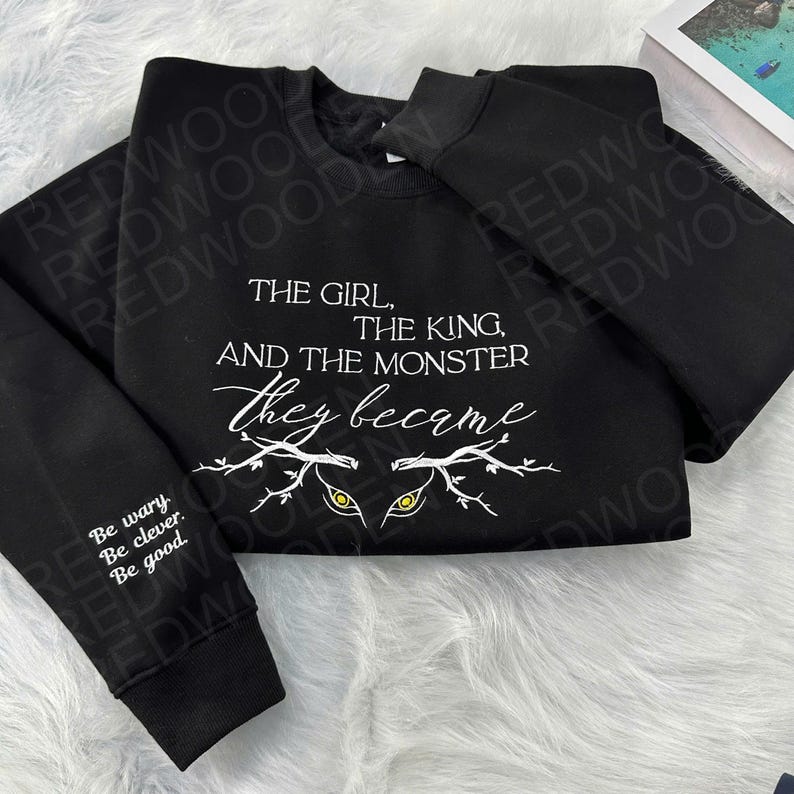 Peut inclure: Sweat-shirt noir avec texte blanc : "THE GIRL, THE KING, AND THE MONSTER, They became." Le design comprend des yeux stylis&eacute;s. La manche gauche porte l'inscription "Be wary. Be clever. Be good."