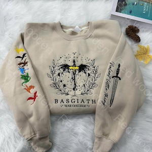Op de afbeelding: Een beige sweatshirt met een zwart-witte afbeelding van een draak met een gele kop en een zwaard op de mouw. De tekst "Basgiath War College" is onder de draak gedrukt. De mouw heeft ook een rij kleurrijke draken.