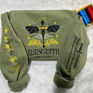 May include: A green sweatshirt with a black dragon embroidered on the front. The dragon is surrounded by yellow clouds and stars. The text "BASGIATH +WAR COLLEGE+" is embroidered below the dragon. The sleeves have a gold embroidered design of a bird with the text "Fly or die".