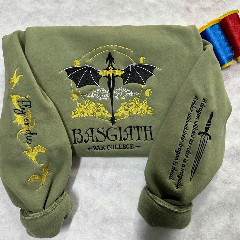 May include: A green sweatshirt with a black dragon embroidered on the front. The dragon is surrounded by yellow clouds and stars. The text "BASGIATH +WAR COLLEGE+" is embroidered below the dragon. The sleeves have a gold embroidered design of a bird with the text "Fly or die".