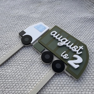 May include: Green and white truck cake topper with the text "august is 2".