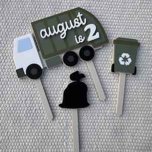 May include: Three green, white, and black cake toppers featuring a garbage truck with the text "august is 2", a trash can with a recycle symbol, and a black garbage bag.