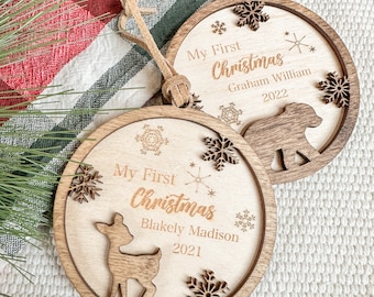 Op de afbeelding: Twee houten kerstversieringen met de tekst "My First Christmas" en namen en data. Eén ornament heeft een hert, de andere een beer, en beide hebben sneeuwvlokuitsparingen. Elke versiering heeft een leren koord om op te hangen.