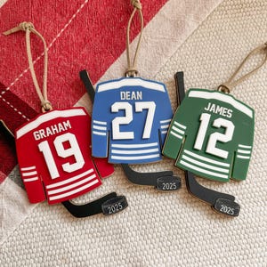 May include: Three hockey jersey ornaments in red, blue, and green, each with a player's name and number. The red jersey has "GRAHAM 19", the blue has "DEAN 27", and the green has "JAMES 12". Each ornament includes a hockey stick and the year "2025".