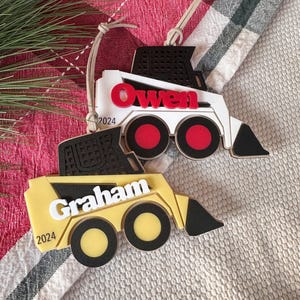 May include: Two wooden toy construction vehicle ornaments, one yellow and one white, with black details. The yellow ornament has the name "Graham" and the year "2024" on it. The white ornament has the name "Owen" on it.