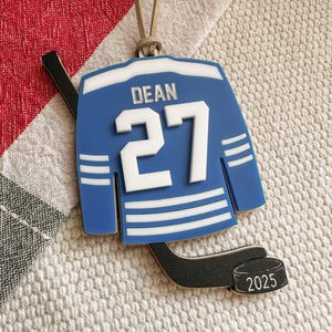 May include: A blue hockey jersey ornament with the name "DEAN" and the number "27" in white. The ornament includes a hockey stick and a puck with the year "2025". It has a tan string for hanging.