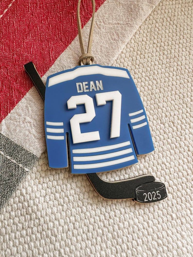May include: A blue hockey jersey ornament with the name "DEAN" and the number "27" in white. The ornament includes a hockey stick and a puck with the year "2025". It has a tan string for hanging.