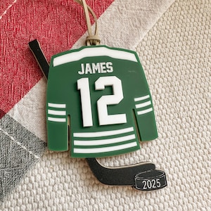 May include: Green and white hockey jersey ornament with the name "JAMES" and the number "12". Includes a black hockey stick and a puck with "2025" printed on it. The ornament has a tan string for hanging.