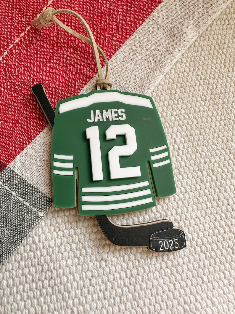 May include: Green and white hockey jersey ornament with the name "JAMES" and the number "12". Includes a black hockey stick and a puck with "2025" printed on it. The ornament has a tan string for hanging.
