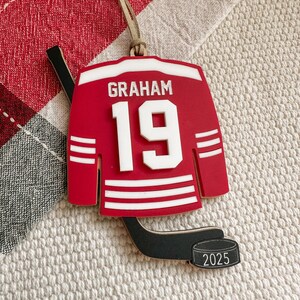 May include: A red hockey jersey ornament with the name "GRAHAM" and the number "19" in white. The ornament includes a hockey stick and a puck with "2025" on it. A tan string is attached for hanging.