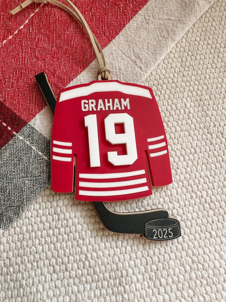 May include: A red hockey jersey ornament with the name "GRAHAM" and the number "19" in white. The ornament includes a hockey stick and a puck with "2025" on it. A tan string is attached for hanging.