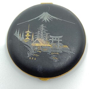 May include: A black round compact with a gold rim and a Japanese scene painted in gold and silver. The scene includes a mountain, a pagoda, a house, a torii gate, and trees. The compact is marked with the number "K24".