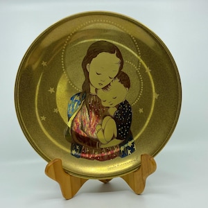 May include: A gold-colored decorative plate featuring an illustration of a mother and child. The plate has a circular design with a star and crescent moon pattern. The mother and child are depicted in warm tones, with the mother wearing a blue and gold garment. The plate is displayed on a wooden stand.