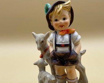 Könnte beinhalten: Eine Porzellanfigur eines Jungen in traditioneller Kleidung mit zwei kleinen Lämmern. Der Junge trägt einen braunen Hut, ein weißes Hemd mit einer roten Schleife und braune Shorts. Die Lämmer sind weiß mit braunen Markierungen.