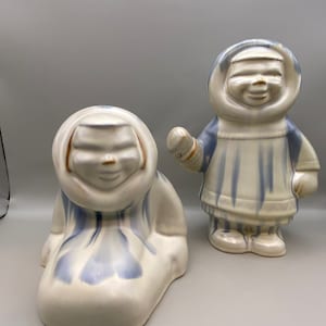 May include: Two ceramic figurines of people wearing blue and white hooded garments. The figurines are standing on a white surface.