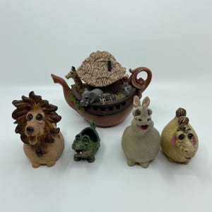 May include: A collection of whimsical animal figurines, including a lion, crocodile, rhino, and a creature with a yellow body and a brown topknot. A brown ark-shaped teapot with a thatched roof and animal figures is also present. The figurines are on a white surface.