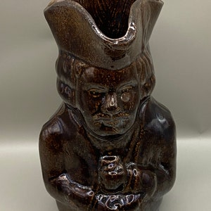 May include: A brown ceramic pitcher shaped like a person wearing a hat. The pitcher has a detailed face and is glazed in a dark brown color.
