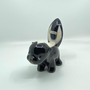May include: A ceramic skunk figurine with a glossy black body and a white stripe down its back. The skunk has a white face with small, detailed features and a curved tail. The figurine is marked with the word "CHEW".