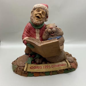 May include: A detailed figurine of a man in a Santa hat and red coat, singing from a book with a squirrel. The base has a banner that reads "CAIRN 1995 CHRISTMAS". The figurine is brown, red, green, and beige.